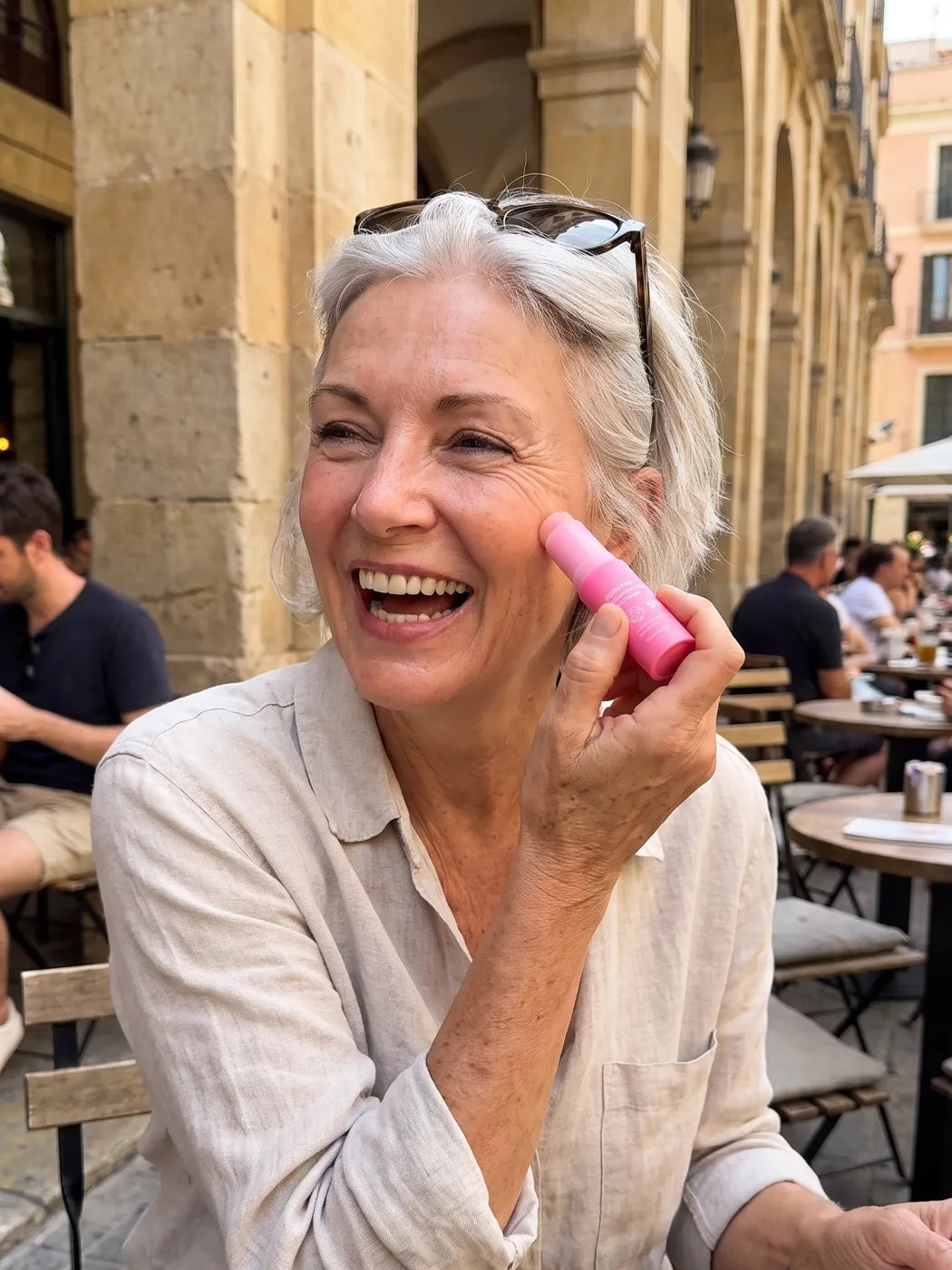 Woman in her sixties laughing at a cafe terrace while applying the Pink Balm to her cheek