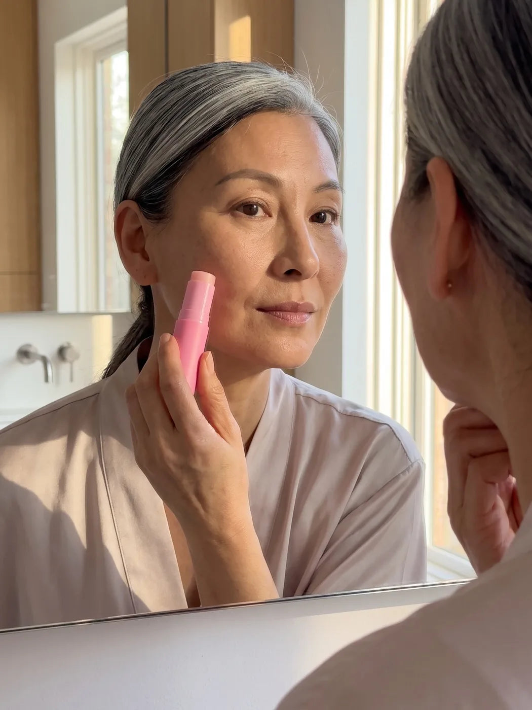 Woman applying the Pink Balm to her cheek in front of a bathroom mirror