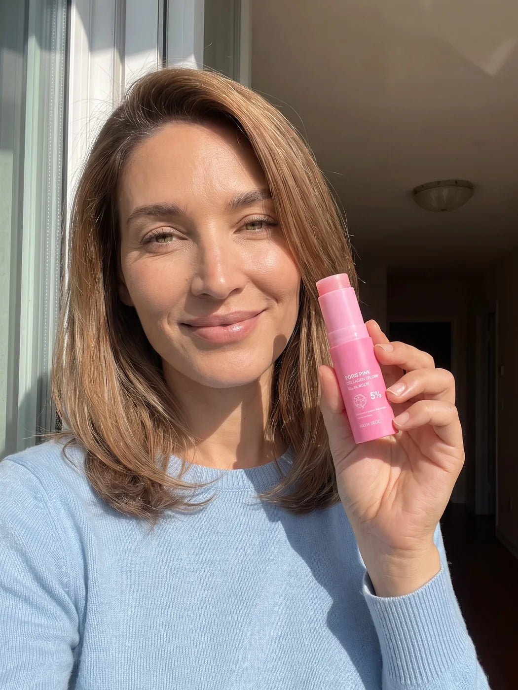 Woman taking a sunlit window selfie, holding the Pink Balm beside her cheek